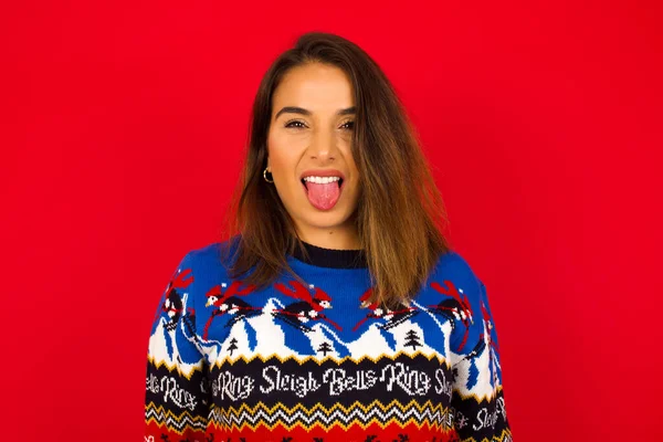 Vücut dili. Kırmızı duvara karşı Noel süveteri giyen, suratını asan, bir şeyden tiksindiğini gösteren, stresli, genç, beyaz bir kadın..