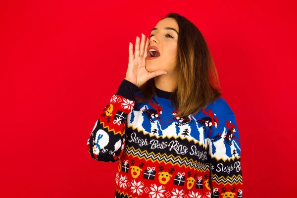 若い美しい白人女性は赤い壁にクリスマスセーターを着て プロフィールビュー 幸せと興奮を探して 叫んで スペースをコピーする呼び出し — ストック写真