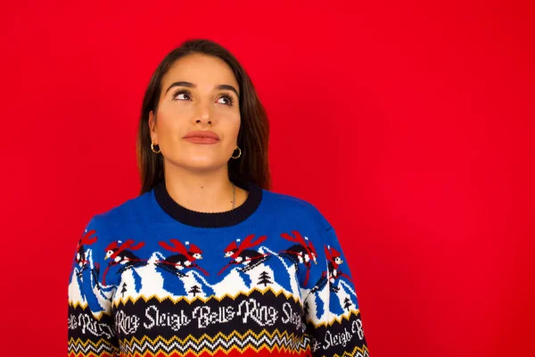 Genç, beyaz bir kadın, kırmızı duvara karşı Noel süveteri giyiyor. Endişeli bir yüzü var. Dudaklarının arasına bakıyor..