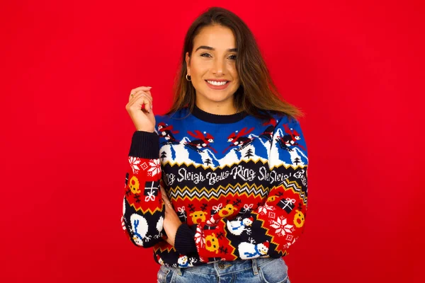 Kırmızı duvara karşı Noel süveteri giyen neşeli, genç, beyaz bir kadın. İyi haberleri dinledikten sonra kameraya mutlu bir ifadeyle bakmak. Kendine güven..