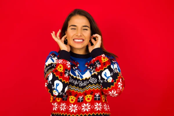 Bu sinir bozucu sesi çıkarmayı kes! Mutsuz, genç, güzel, beyaz bir kadın Noel süveterini kırmızı duvara dayamış, endişeli bir yüz ifadesi takınıyor, kulaklarını parmaklarıyla tıkıyor, gürültüden rahatsız oluyor..