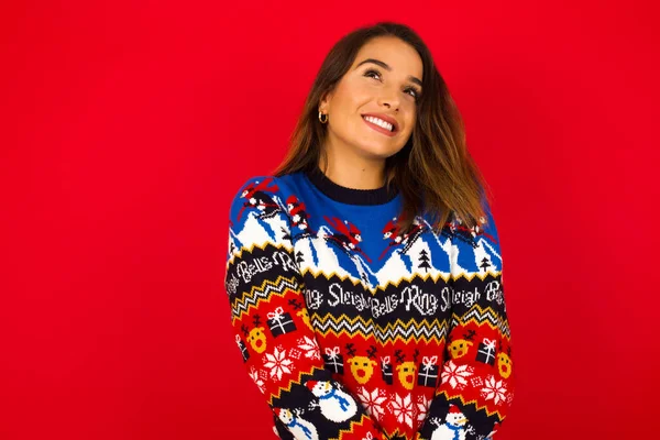 Kırmızı duvara karşı Noel süveteri giyen gizemli, güzel beyaz kadın portresi, gizemli bir gülümsemeyle yukarı bakıyor. Reklam kavramı.