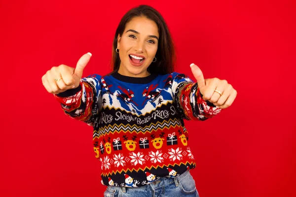 赤い壁にクリスマスのセーターを着て 親指でポジティブなジェスチャーを笑顔で幸せにします カメラを見て 勝者のジェスチャー — ストック写真
