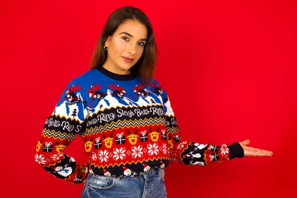 歓迎のジェスチャーで腕で赤い壁に対してクリスマスのセーターを着用する慎重な女性の肖像画 — ストック写真