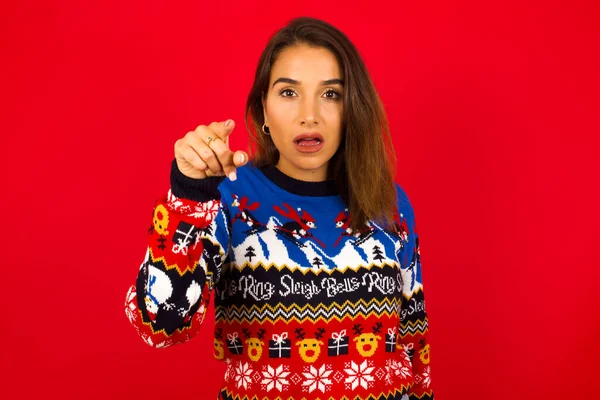 Kafkasyalı bir kadın kırmızı duvara karşı süveter giyip kameraya işaret parmağıyla işaret ediyor. Sürpriz ve reklam konsepti.