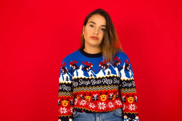 Kasvetli, sıkılmış genç, beyaz bir kadın Noel süveterini kırmızı duvara dayamış kaşlarını çatmış, yukarı bakıyor, çok fazla konuşmaktan rahatsız oluyor, yorgun ve gitmek istiyor..