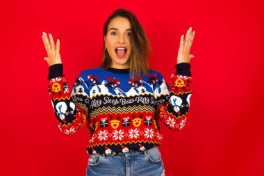 Neşeli, şanslı, genç, beyaz bir kadın Noel süveterini kırmızı duvara dayamış, tezahürat yapıyor, başarıyı kutluyor, yumruklarını sıkarak evet diye bağırıyor.