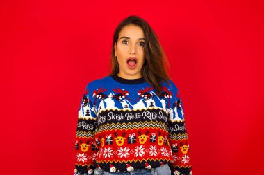 Duygusal olarak çekici genç beyaz bir kadın kırmızı duvara karşı Noel süveteri giyiyor, ağzı açık, büyük bir şaşkınlık ve korku ifade ediyor, kameraya bakıyor. Beklenmedik haberler ve insani tepkiler.