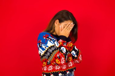 Üzgün, genç, beyaz, güzel bir kadın Noel süveteri giyiyor, yüzünü ellerle kaplıyor ve ağlıyor..