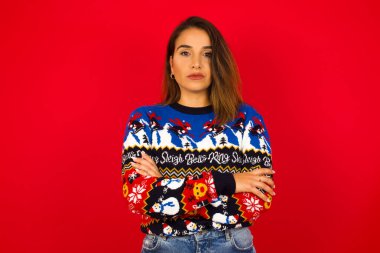 Kızgın genç beyaz bir kadının Noel süveterini kırmızı duvara dayayıp kameraya baktığı bir resim..