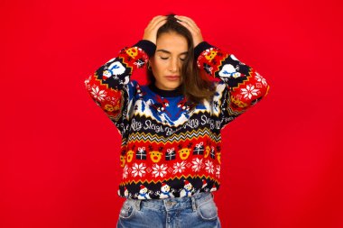 Kızıl duvara karşı Noel süveteri giyen, elleriyle başını tutan, şiddetli baş ağrısından muzdarip, şakaklara parmaklarını bastıran güzel beyaz bir kadın.