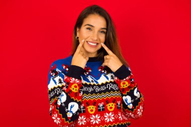 Güçlü, sağlıklı, beyaz dişler. Kırmızı duvara dayalı Noel süveteri giyen mutlu beyaz kadın portresi. Parıldayan gülümseme kusursuz beyaz dişleri işaret ediyor..