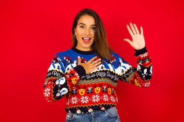 Yemin ederim, pişman olmayacağına söz ver. Samimi, genç, beyaz bir kadının portresi Noel süveterini kırmızı duvara dayamış, bir kolunu kaldırıp kalbe dokunmuş yemin ediyor, doğruyu söylüyor, inanmanızı istiyorum..