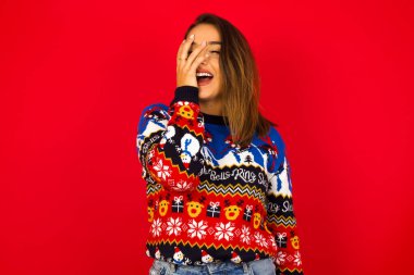 Karizmatik, kaygısız, neşeli, güzel, beyaz bir kadın Noel süveterini kırmızı duvara dayamış, yüksek sesle gülmeyi seviyor. Duygularını gizlemiyor, kıkırdıyor..