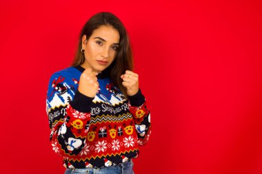 Çekici genç beyaz kadın portresi. Kırmızı duvara karşı Noel süveteri giyiyor. Boks pozisyonunda önünde el ele tutuşuyor..