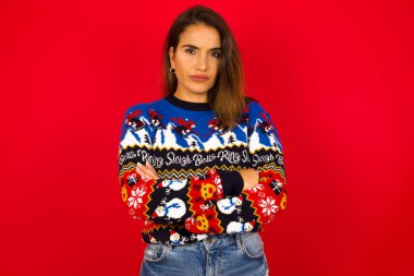 Kızgın, genç, beyaz bir kadının Noel süveterini kırmızı duvara dayayıp kollarını kavuşturduğu bir resim. Hayal kırıklığına uğramış bir ifadeyle kameraya bakmak.