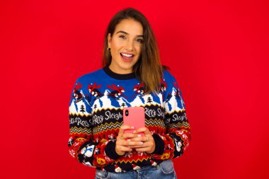 Gülümseyen genç beyaz kadın, kırmızı duvara karşı Noel süveteri giyiyor, dost canlısı ve mutlu bir şekilde cep telefonuyla aynada selfie çekiyor..