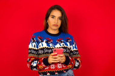 Kafası karışmış genç, beyaz bir kadının portresi. Kırmızı duvara karşı Noel süveteri giyiyor, cep telefonu tutuyor, omuzlarını silkiyor ve kaşlarını çatıyor..