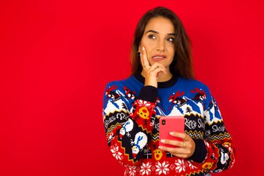 Düşünen genç beyaz bir kadının Noel süveterini kırmızı duvara dayayıp cep telefonu kullandığını ve el ele tutuştuğunu hayal et. Karar almak ve sosyal medya kavramı.