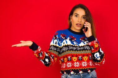 Kızıl duvara karşı Noel kazağı giyen genç, güzel beyaz bir kadın, telefonda konuşurken yüz yüze, utanç ve şaşkınlıktan şok olmuş, sinirli ve hayal kırıklığına uğramış. Hata için korku ve üzüntü.