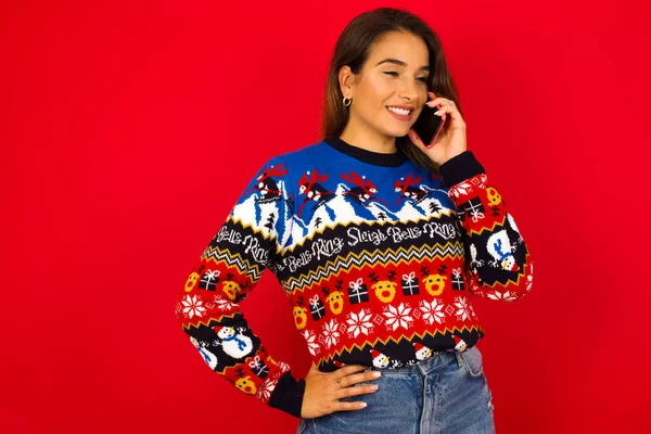 成功快乐的年轻美丽的高加索女人穿着圣诞毛衣靠着红墙 与朋友在手机上交谈的画像 生活方式和交流概念 — 图库照片