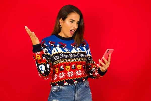 Photo of outraged annoyed Young beautiful Caucasian woman wearing Christmas sweater against red wall,  holds cell phone, makes call, argues with colleague,  expresses negative emotions. People and anger.