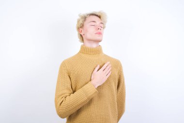 Tatmin olmuş, gülümseyen, beyaz arka planda duran yakışıklı, beyaz tenli, sarışın bir adam ellerini karnının üstüne koyuyor. Lezzetli bir yemek yedikten sonra iyi bir ruh hali içinde olması tok olduğunu gösteriyor. Midemde hoş bir his var..