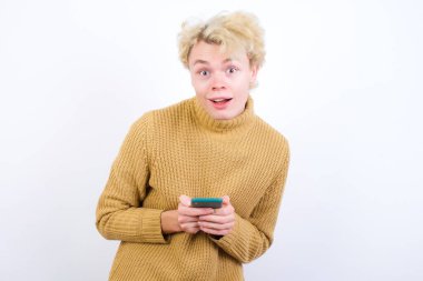 Gülümseyen yakışıklı beyaz sarışın adam beyaz arka planda duruyor. Arkadaş canlısı ve mutlu bir şekilde cep telefonu tutuyor. Aynada selfie çekiyor..