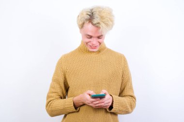 Gülümseyen yakışıklı, beyaz arka planda duran beyaz saçlı, cep telefonu kullanan, mesajlaşan, arkadaşlarıyla mesajlaşan, akıllı telefona bakan beyaz bir adam. Modern teknolojiler ve iletişim.