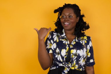 Omg konsepti. Şaşırmış güzel afro-amerikalı kadın, şaşırmış bir ifadeyle gözlük takıyor, gözlerini ve ağzını iyice açıyor, başparmağıyla işaret ediyor, garip bir şey gösteriyor. 