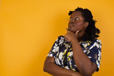Düşünceli gülümseyen Afrikalı Amerikalı kadın portresi elini çenesinin altında tutuyor, yan bakıyor, ilgi çekici bir şey düşünüyor ya da merak ediyor, sıradan giyiniyor, gri stüdyoya karşı poz veriyor. Karar alma kavramı.