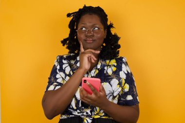Düşünen, güzel, Afro-Amerikan bir kadının parlak duvar arkasında cep telefonuyla izole edilmiş bir şekilde poz vermesi..