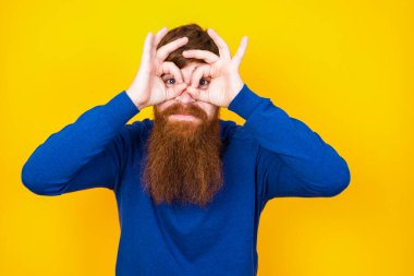 Neşeli, yakışıklı, kızıl saçlı, beyaz adam sarı duvarın önünde duruyor. Tamam işareti iki gözünün üstünde, gözlük takıyormuş gibi yapıyor..