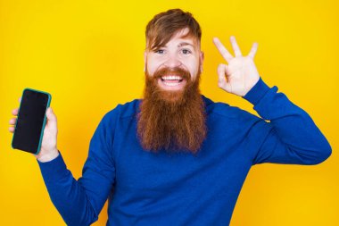 Heyecanlı, kızıl sakallı beyaz adam sarı duvarın karşısında duruyor. Akıllı telefon ekranı açık, göz kırpıyor ve el işareti yapıyor. Reklam kavramı.