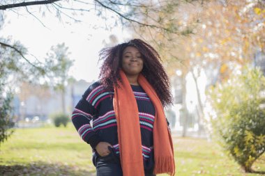 Genç, güzel, gülümseyen Afrikalı Amerikalı kadın sonbahar parkında kazak ve turuncu eşarp giyiyor.   