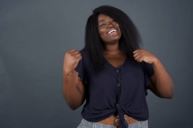 Gri arka planda poz veren Afro-Amerikalı kadın, başarısını ve zaferini sevinçle kutluyor ve yumruklarını zevkle sıkıyor. Tüylü, şanslı kadın hedefine ve hedeflerine ulaştığına seviniyor. Olumlu duygular, duygular.