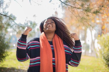 Genç, güzel, mutlu, Afro-Amerikan kadın süveter ve turuncu fular giyerek sonbahar parkında yumruk kaldırıyor.  