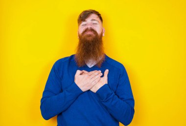 Sarı duvara karşı duran kızıl saçlı beyaz adam gözlerini kapatır ve ellerini kalbine yakın tutarak samimi duygular ifade eder, iyi kalpli ve dürüst davranır. Vücut dili ve gerçek duygular kavramı.