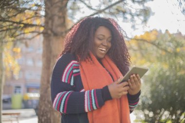 Güz parkında kazak ve eşarp giyen güzel Afro-Amerikan kadın elinde tablet bilgisayarla geziyor.   