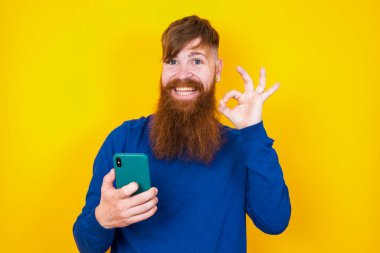Mutlu, kızıl saçlı, sakallı beyaz adam sarı duvara karşı duruyor. Akıllı telefonuyla mesaj yolluyor ya da selfie çekiyor ve eliyle imzasını atıyor..