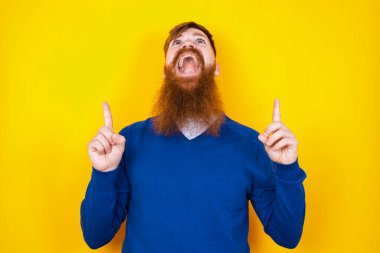 Yakışıklı kızıl sakallı beyaz adam, sarı duvara karşı duruyor, olumlu bir ifadeyle, her iki işaret parmağıyla işaret ediyor, geniş bir gülümsemesi var. Şuraya bakın, lütfen..