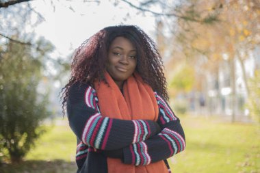 Güz parkında kolları çaprazlanmış kazak ve eşarp giyen güzel Afro-Amerikan bir kadın.  