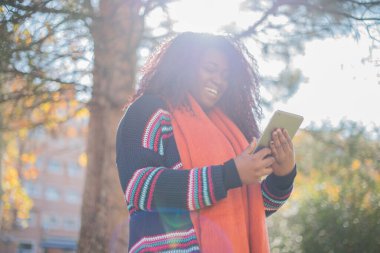 Genç ve güzel Afro-Amerikalı kadın sonbahar parkında tablet bilgisayar kullanarak kazak ve eşarp giyiyor.  