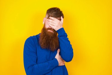 Yakışıklı kızıl sakallı beyaz adam, sarı duvarın karşısında durup, aptalca bir duruma hayret içinde gülümserken fotoepalm hareketi yapıyor..