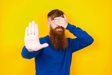 Sarı duvara karşı duran kızıl sakallı beyaz adam, gözleri avuç içiyle kaplıyor ve dur hareketi yapıyor, saklanmaya çalışıyor. Bana bakma, görmek istemiyorum, utanıyorum ya da korkuyorum..