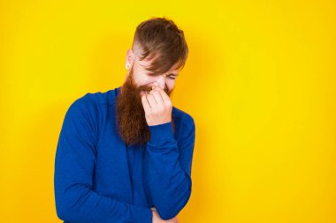 Yakışıklı kızıl sakallı beyaz adam kötü bir koku yüzünden sarı duvarın önünde dikilip burnunu tutuyor..