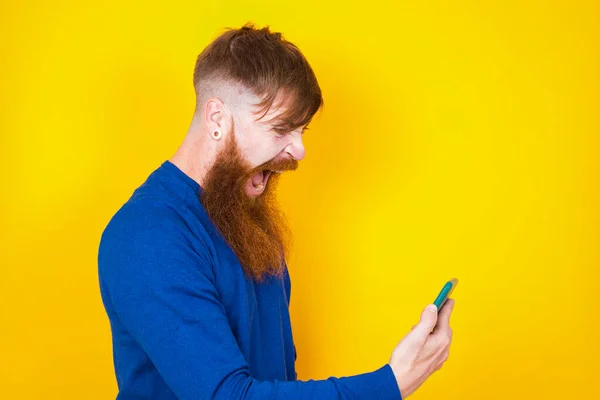 Photo of outraged annoyed handsome red haired bearded Caucasian man standing against yellow wall holds cell phone, makes call, argues with colleague,  expresses negative emotions. People and anger.