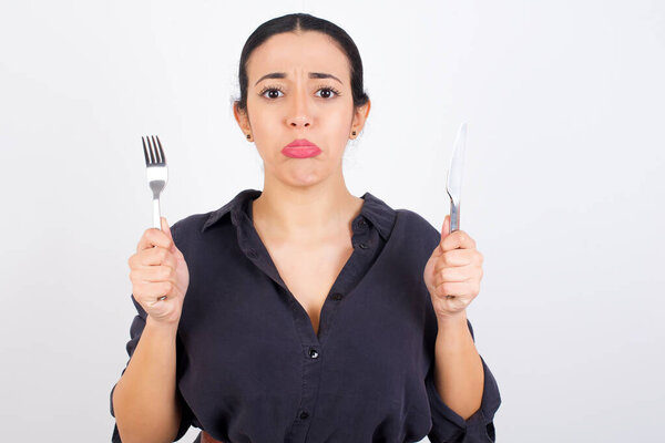 Молодая красивая грустная арабская женщина в платье на белом фоне студии с вилкой и ножом в студии 