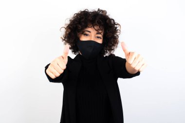  Tıbbi maske takan beyaz bir kadın olumlu bir jest yapıyor. Başparmağıyla gülümsüyor ve başarı için seviniyor. Kameraya bak, kazanan jest..