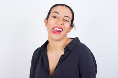 Genç, güzel bir Arap kadın beyaz stüdyo geçmişine karşı gri elbiseler giyiyor mutlu ve komik bir yüzü var gülümsüyor ve dil gösteriyor..